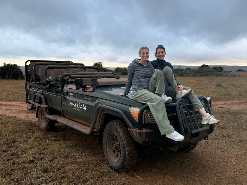 Zwei Vögele-Mitarbeiterinnen sitzen bei bewölktem Himmel auf dem Safari-Fahrzeug in der Steppe des Amakhala Game Reserves in Südafrika.