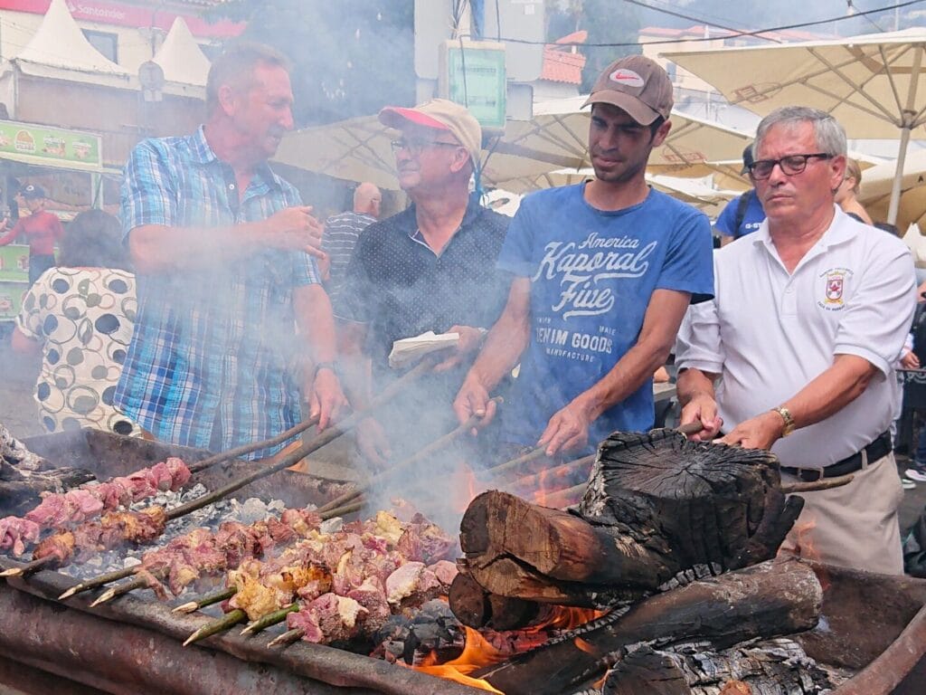 Vier Männer stehen an einem Holzkohle-Grill und grillieren Fleischstücke an einem Holzspiess